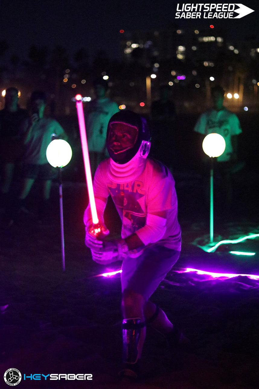 A person in a helmet and glowing white outfit holds a pink lightsaber-style stick at night outdoors, with other participants and neon light markers visible in the background. Visible logos include Lightspeed Saber League and HeySabre.