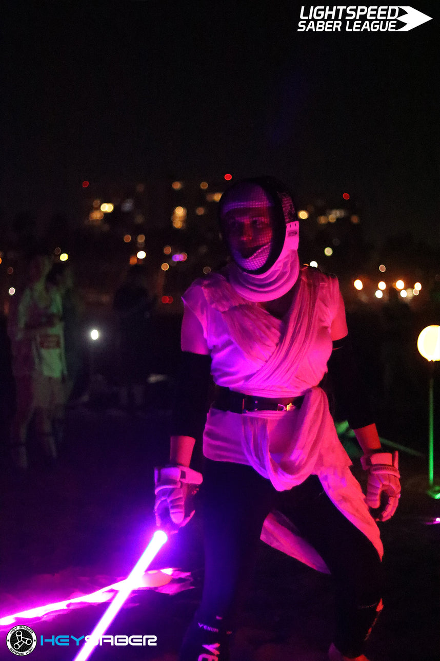 A person in a white and black costume holding a glowing pink lightsaber at night, with blurred lights and other event lights in the background. The 'LIGHTSPEED SABER LEAGUE' logo is in the top right, and 'HEY SABER' is in the bottom left.