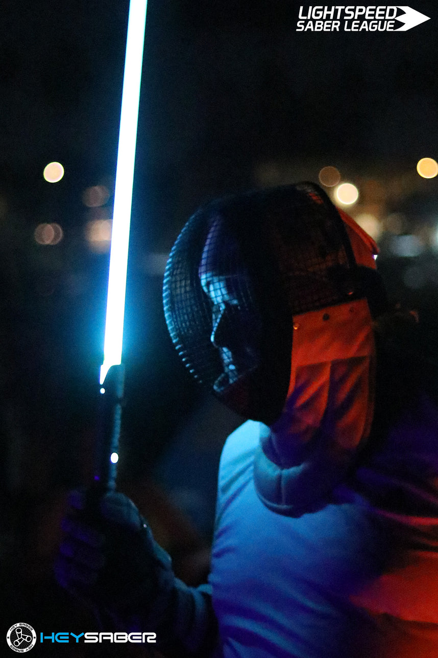 A fencer in protective gear holding a glowing blue lightsaber at night, with the Lightspeed Saber League logo in the top right and a HEGY SABER logo in the bottom left.